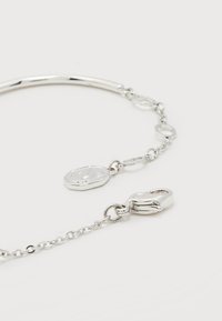 Ασημένιο βραχιόλι με αλυσίδα, κούμπωμα σε σχήμα νύχι αστακού και ένα μικρό οβάλ μενταγιόν που φέρει χαραγμένο το γράμμα "J" σε λευκό φόντο.