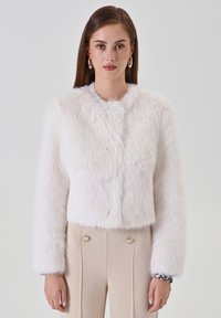 Weiße, kurz geschnittene Jacke aus Kunstfell mit langen Ärmeln, rundem Ausschnitt und weicher Textur. Ergänzt durch beige, hochtaillierte Hose mit goldenen Akzenten.