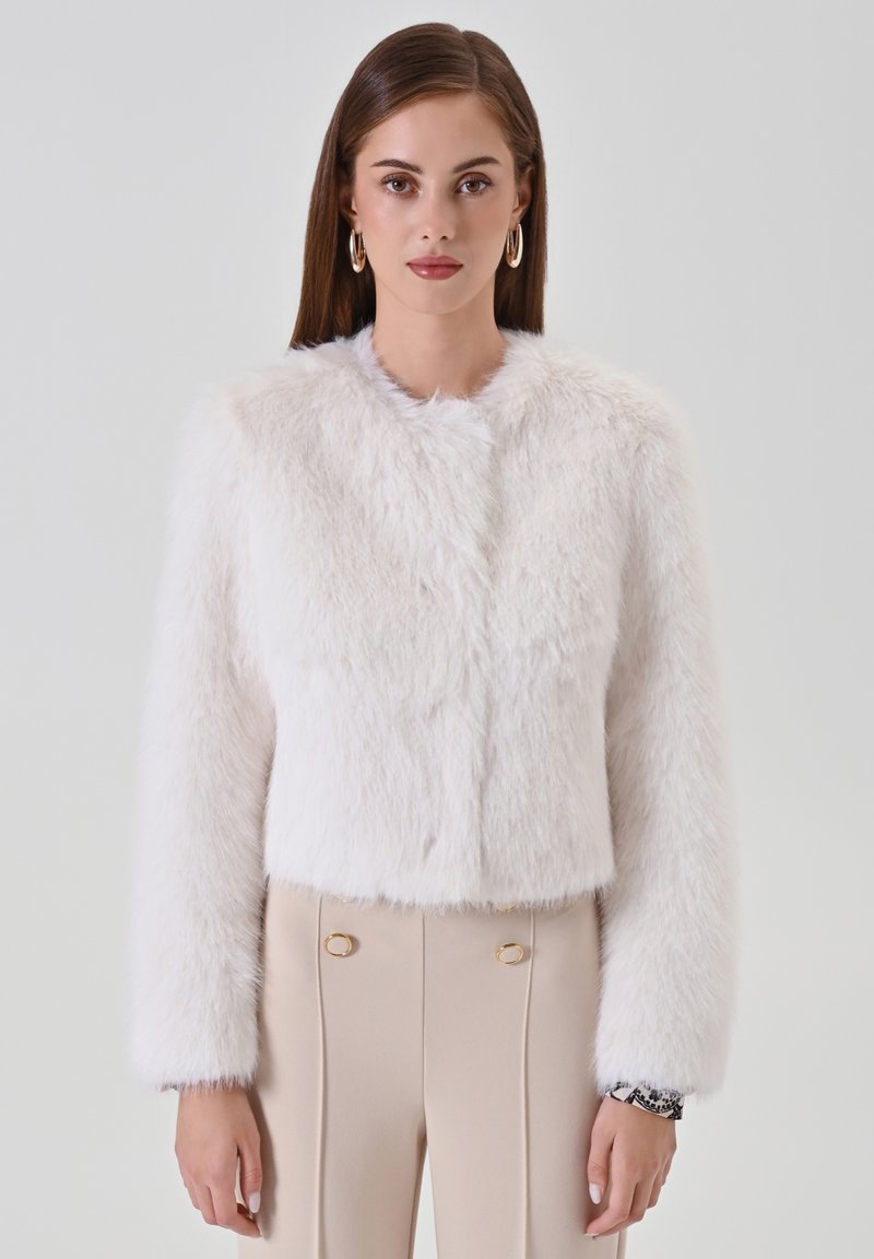 Weiße, kurz geschnittene Jacke aus Kunstfell mit langen Ärmeln, rundem Ausschnitt und weicher Textur. Ergänzt durch beige, hochtaillierte Hose mit goldenen Akzenten.