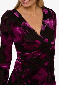 Žena v černých šatech s živými fialovými sametovými květinovými vzory a výstřihem do V, usmívající se s dlouhými blond vlasy na viděné.
