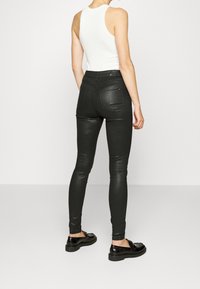 Jeans skinny negros de talle alto con acabado brillante, que presentan dos bolsillos traseros y una altura media, combinados con una camiseta sin mangas blanca y zapatos negros.
