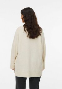 Beżowy, oversize sweter z opadającymi ramionami, bocznymi rozcięciami i miękką fakturą, zestawiony z ciemnymi spodniami. Widok z tyłu.