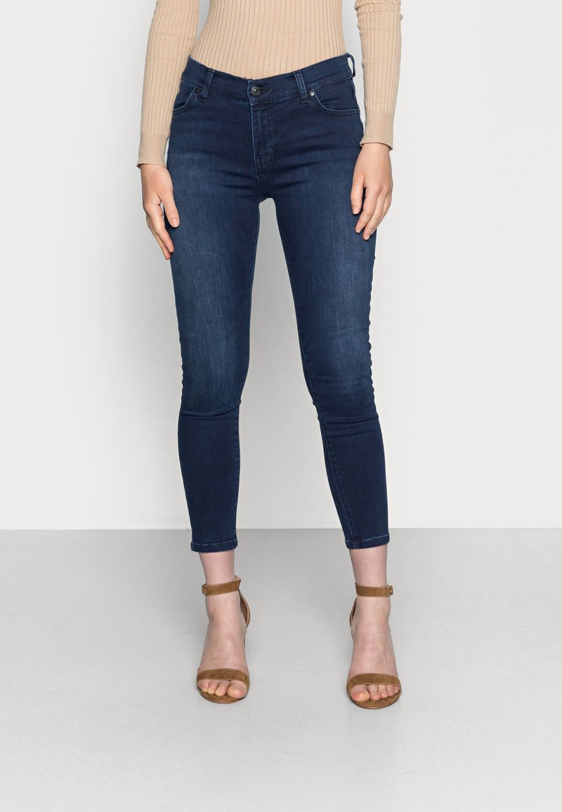 Jeans skinny de color azul oscuro, confeccionados en denim, con un diseño ajustado y corte a la altura del tobillo, combinados con sandalias marrones de tiras.