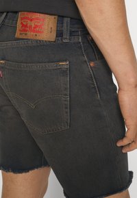 Homme portant un short en denim noir Levi’s avec des ourlets effilochés, des coutures visibles sur la poche arrière et un écusson en cuir marron sur la ceinture.