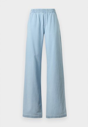 Femme debout, une main dans la poche, portant un haut sans manches crème, un pantalon large bleu clair et des chaussures foncées sur fond blanc.