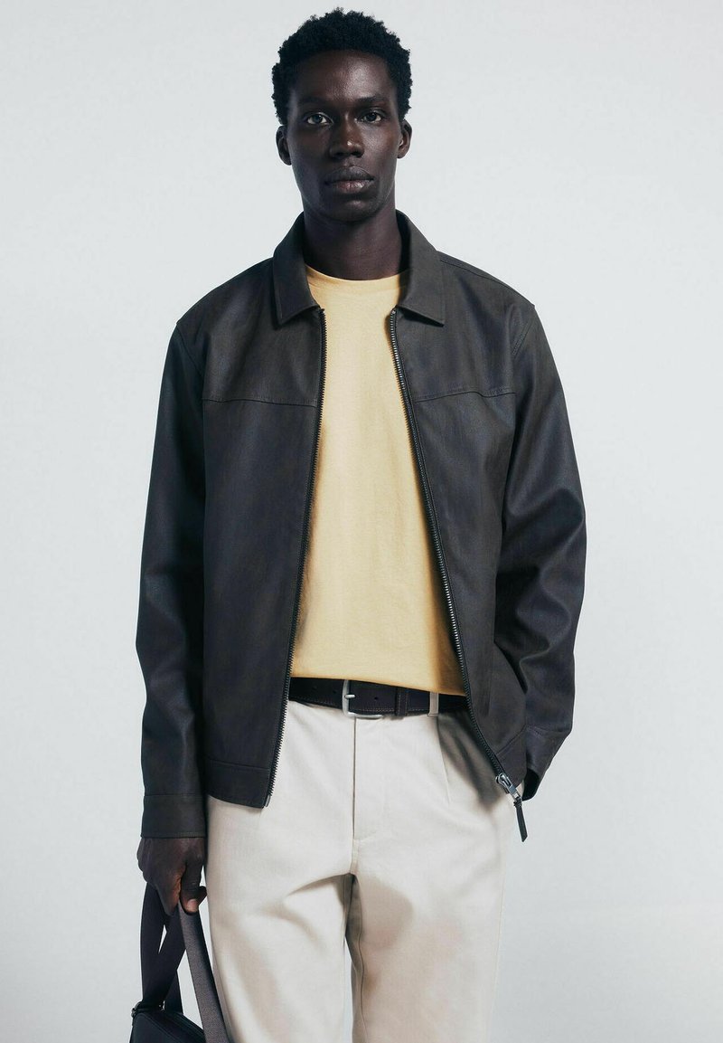 Giacca di pelle marrone con colletto, chiusura con zip e due tasche frontali, indossata sopra una maglietta gialla e pantaloni beige chiaro.