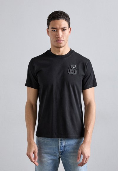 T-shirt noir en coton avec un col rond, des manches courtes, présentant un logo de serpent texturé sur la zone gauche de la poitrine.