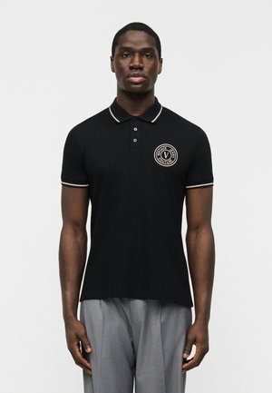 Férfi fekete galléros pólóban, fehér szegéllyel és a bal mellkason egy kör alakú, "Versace Jeans Couture" logóval, szürke nadrággal párosítva.