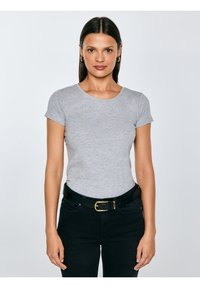 Młoda kobieta o prostych ciemnych włosach, nosząca złote kołnierzowe kolczyki, dopasowaną szarą koszulkę z krótkim rękawem oraz czarne spodnie z paskiem, stojąca.