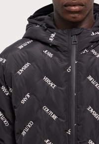 Juodas pūkinis švarkas su kapišonu, su baltu siuvinėjimu „Versace“ ir „Jeans“ įvairiomis orientacijomis, užtrauktuku ir tekstūruotu paviršiumi.