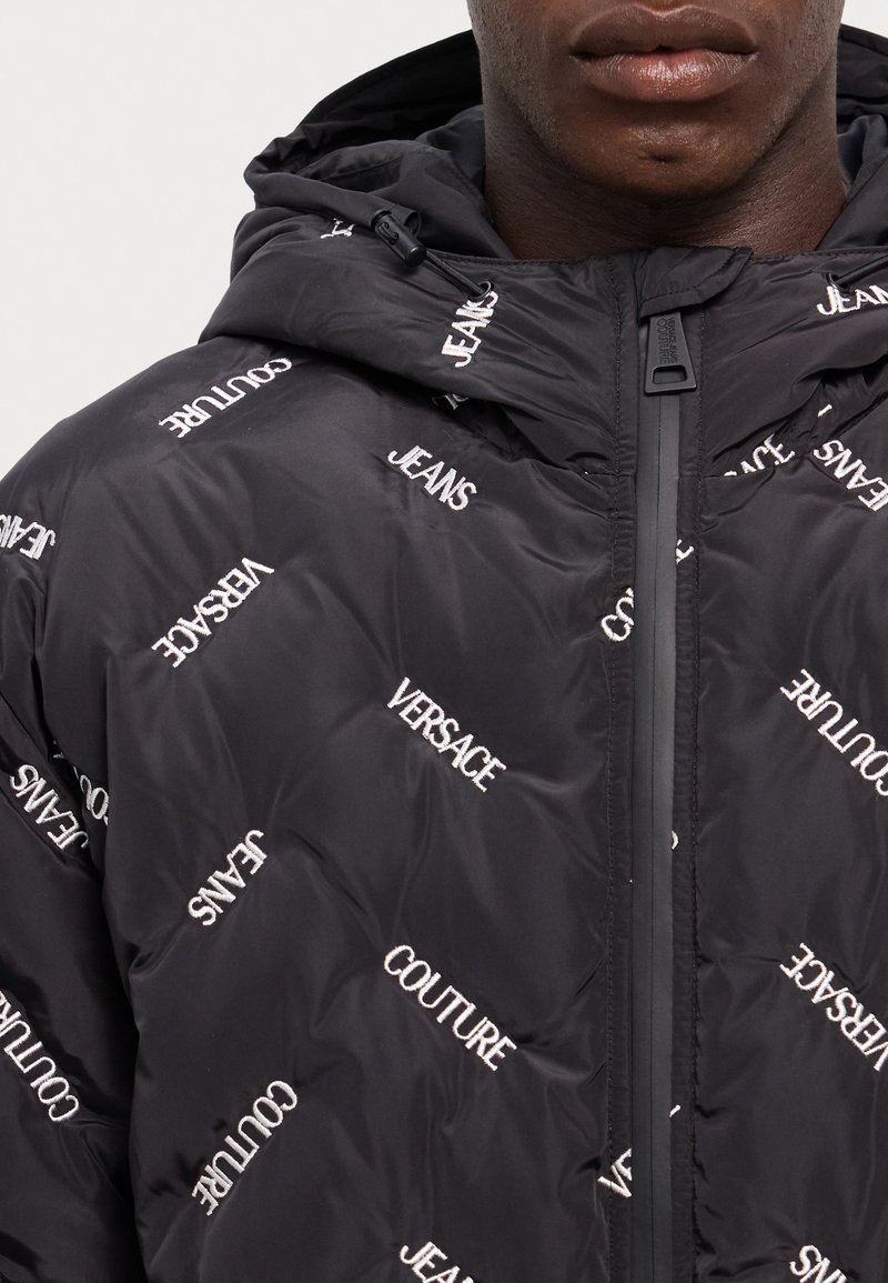 Chaqueta acolchada negra con capucha, que presenta el texto bordado en blanco "Versace" y "Jeans" en varias orientaciones, cierre con cremallera y acabado texturizado.