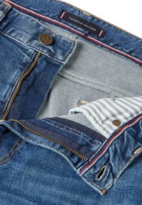Jean en denim bleu non fermé avec fermeture éclair visible, bouton gravé du nom de la marque, doublure intérieure rayée de la poche et étiquette Tommy Hilfiger à l'intérieur de la ceinture.