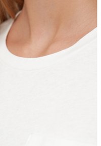 Λευκό βαμβακερό μπλουζάκι με στρογγυλή λαιμόκοψη, λείο ύφασμα και διακριτικές λεπτομέρειες ραφής στην περιοχή του γιακά και των ώμων.