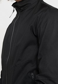 Svart jacka med hög krage, med en hel dragkedja och dragkedjefickor på sidorna. Tyget har en slät, matt yta.