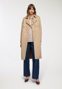 Cappotto beige in suede con un grande colletto, due tasche frontali, di lunghezza media, indossato sopra una camicia chiara e abbinato a jeans in denim a gamba larga.