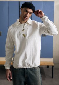 Bílý svetr s límcem, který má žlutou nášivku lva a logo Adidas. Rukávy jsou zakončeny manžetami a má stahovací lem.