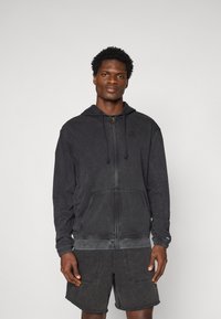 Schwarze Zip-Hoodie mit Kapuze und Kordelzug, Taschen und gerippten Bündchen, aus einem weichen, strukturierten Stoff; getragen mit dunklen Shorts.