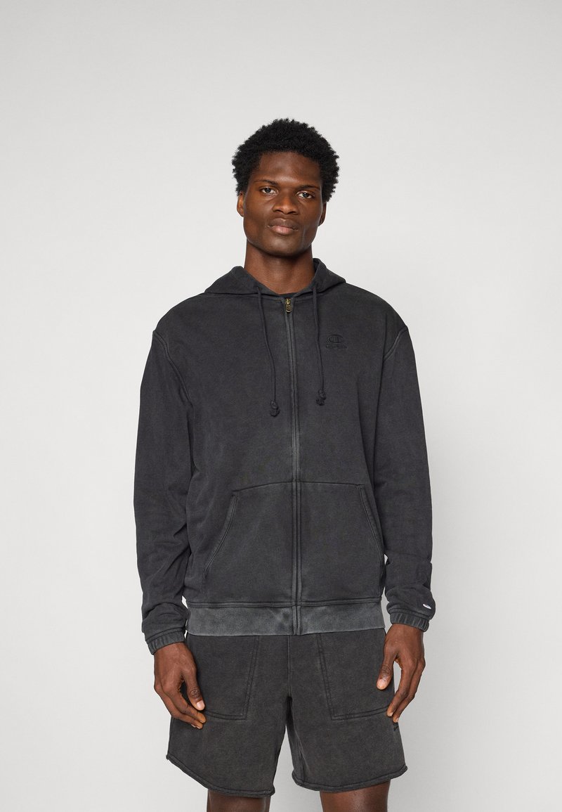 Schwarze Zip-Hoodie mit Kapuze und Kordelzug, Taschen und gerippten Bündchen, aus einem weichen, strukturierten Stoff; getragen mit dunklen Shorts.