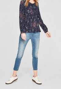 Blusa a maniche lunghe floreale blu navy con polsini aderenti, abbinata a jeans azzurri alla caviglia e sneakers bianche con suola marrone.
