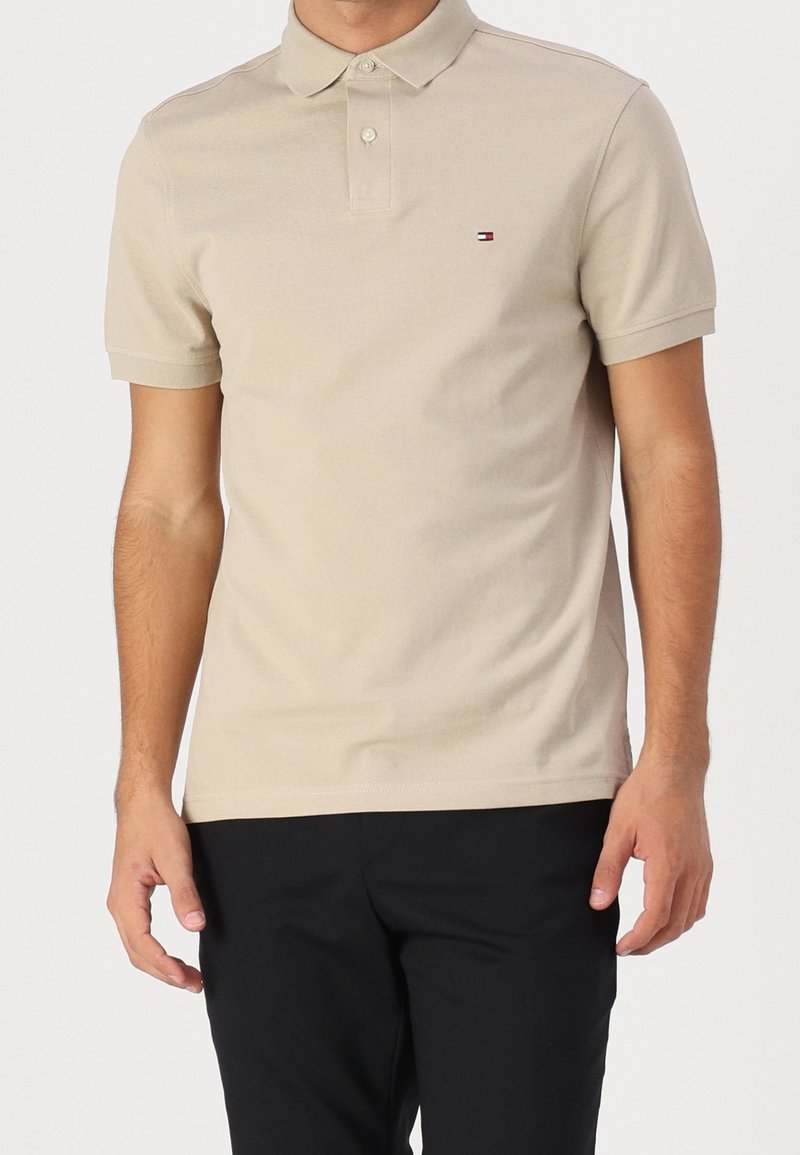 Beige polo tričko s límečkem a krátkými rukávy. Obsahuje malé logo na levé straně hrudi. Tkanina vypadá hladce a texturovaně.