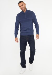Marinblauer Reißverschluss-Pullover mit gerippter Textur über der Brust, kombiniert mit dunklen Jeans und weißen Sneakern. Glattes Strickmaterial, lässige Passform.