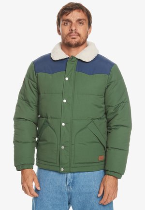 Green quilted jacket with navy blue shoulder yoke, white furry collar, snap buttons, and two front pockets. Worn over blue jeans.