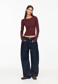 Langärmliges burgunderfarbenes Oberteil mit figurbetontem Schnitt, kombiniert mit locker sitzenden, dunkelblauen Jeans. Das Outfit zeichnet sich durch einen lässigen Stil und klassische Schnitte aus.