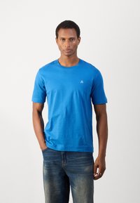 T-shirt azul de manga curta feito de tecido suave, com um decote redondo e um pequeno logótipo branco no lado esquerdo do peito. Usado com jeans de ganga.