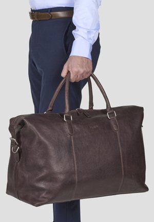 Bruine leren sporttas met een gladde textuur, dubbele handvatten, zilveren hardware en een ritsluiting; heeft zichtbare stiksels en een logo.