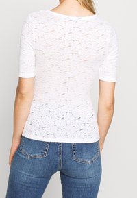 Bílý krajkový top s krátkými rukávy, s květinovým vzorem a mírně průhlednou texturou. Ukončeno kulatým výstřihem a úzkým střihem.