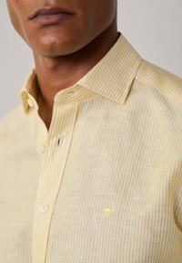Camicia a righe giallo chiaro con colletto a punta, caratterizzata da una leggera texture e un piccolo logo ricamato nell'angolo in basso a destra.