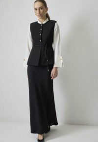 Černá peplum vesta se zlatými knoflíky přes bílou košili, doplněná obepínacími černými maxi sukněmi. Hladká tkanina, jemná textura, délka ke kotníkům.