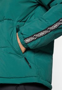 Τζάκετ σε πετρόλ χρώμα με επένδυση, με μαύρη και λευκή ταινία με σχέδιο στο μανίκι, χέρι σε φερμουάρ πλαινής τσέπης, ελαστικό μανίκι ορατό στον καρπό.