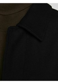 Svart ullmixkappa med en framträdande krage, slät yta och en strukturerad design, delvis överlappande en brun tyg under.