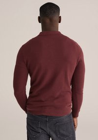 Bordo pulover džemper s teksturiranim pletenjem, krojem prilagođenim tijelu i rebrastim rubom. Uparen s tamnim trapericama prikazanim na neutralnoj pozadini.