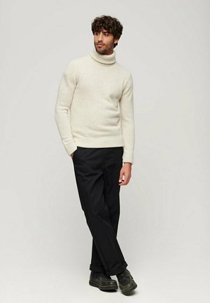 Suéter de cuello alto en color crema, hecho de un material suave, combinado con pantalones negros y zapatos oscuros. Diseño simple con bordes acanalados y una silueta ajustada.