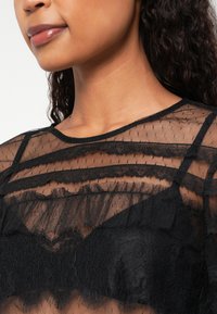 Black sheer top with layered striped design, featuring lace accents and a round neckline, showcasing a black lacy undergarment underneath.