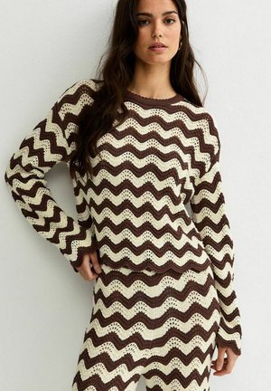 Donna che indossa un maglione a maglia marrone e crema con motivo a zigzag e una gonna abbinata, in piedi contro un muro bianco semplice.