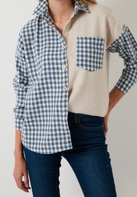 Camicia a quadri blu e bianchi con un lato in beige solido. Presenta una chiusura con bottoni frontale, una tasca e un colletto a contrasto.