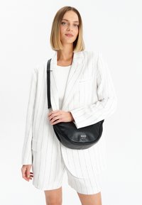 Borsa a tracolla in pelle nera con una forma curva, dotata di un design compatto e di un accento con logo. Il modello indossa una giacca a righe bianche e pantaloni corti.