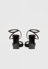 Μαύρα πέδιλα με ψηλό τακούνι και γυαλιστερό φινίρισμα, σχέδιο με ανοιχτή μύτη και λεπτά σταυρωτά λουράκια στον αστράγαλο. Τετράγωνα χοντρά τακούνια προσφέρουν σταθερότητα.