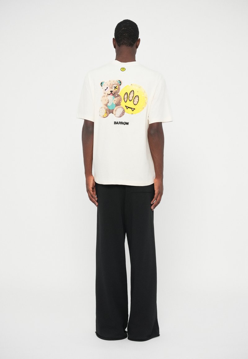 Cream cotton t-shirt featuring a colorful graphic of a bear and smiley face on the back, paired with black wide-leg trousers.