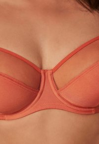 Oranje bh met een gladde stof, doorschijnend gaasontwerp bij de cups en een centrale naad die een gestructureerde vorm creëert.