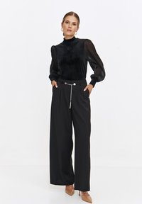 Czarna bluzka z długim rękawem i wysokim kołnierzem, zestawiona z szerokimi czarnymi spodniami i beżowymi obcasami; posiada detal w postaci łańcuszkowego pasa.