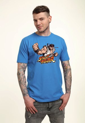 Mand med tatoveringer modellerer en blå Street Fighter T-shirt med en kæmperfigur i slagposition på forsiden.