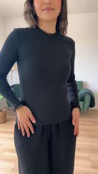 Top preto de malha com mangas longas, com um decote frisado e um leve detalhe de frufru nas mangas, combinado com calças pretas.