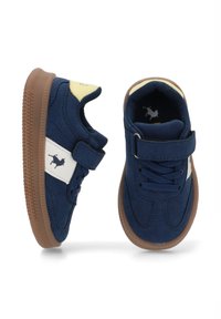 Zapatillas de deporte azul marino con material de ante, panel lateral blanco, correa de Velcro y suela de goma marrón, presentando un pequeño logo en el lateral.