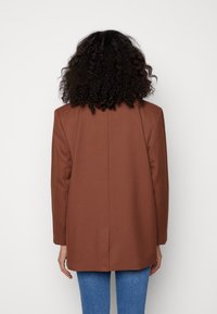 Mujer con pelo rizado, vestida con un blazer marrón y vaqueros azules, vista desde atrás sobre un fondo blanco liso.