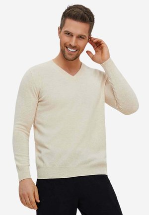 Lächelnder Mann trägt einen cremefarbenen V-Ausschnittpullover und schwarze Hose, mit einer Hand, die sein Ohr vor neutralem Hintergrund berührt.