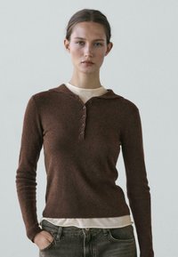 Young woman with light skin and pulled-back hair wears a brown long-sleeve knit top over a beige shirt, posing with one hand in jean pocket.